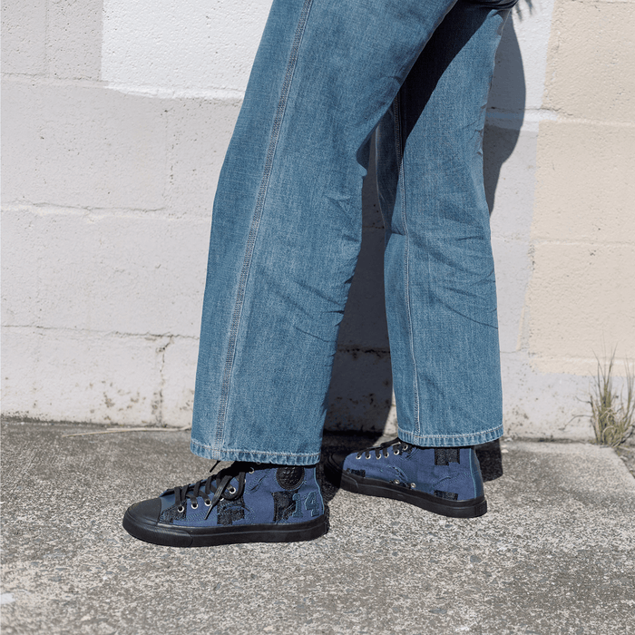 Person wearing blue jeans and matching sneakers standing on a concrete surface with a light-colored wall in the background.