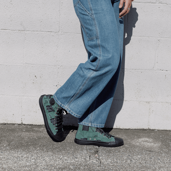 Person wearing blue jeans and green patch work sneakers against a white wall.