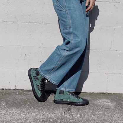 Person wearing blue jeans and green patch work sneakers against a white wall.