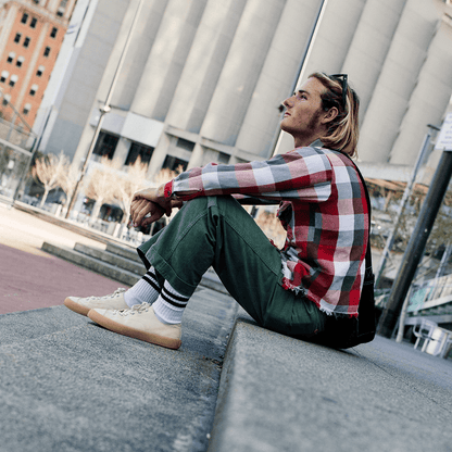 Person sitting on a concrete ledge wearing a plaid shirt, green pants, and beige low-top sneakers in an urban setting.