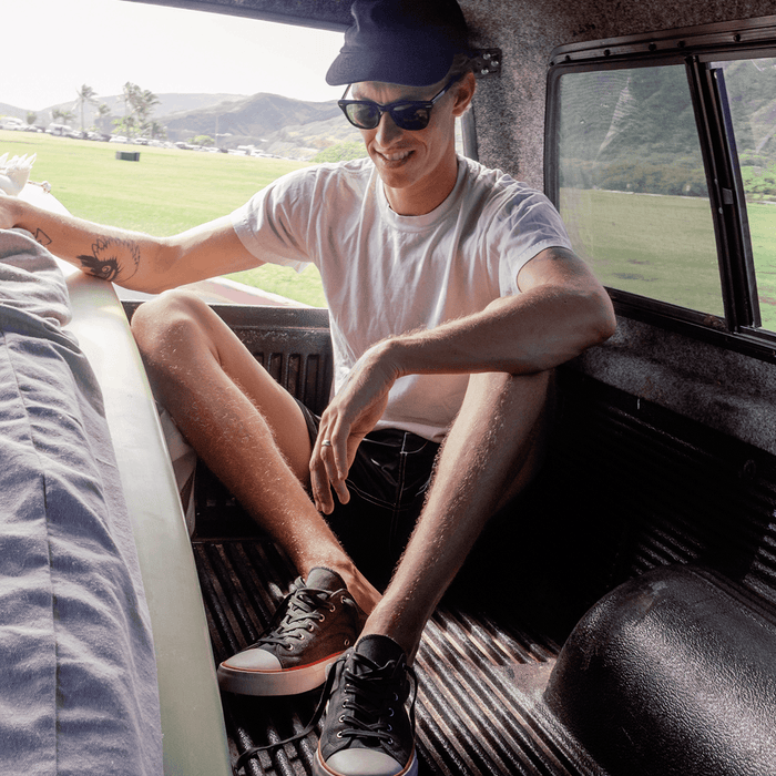 Man wearing sunglasses, black low-top sneakers and shorts sitting in the back of an SUV.