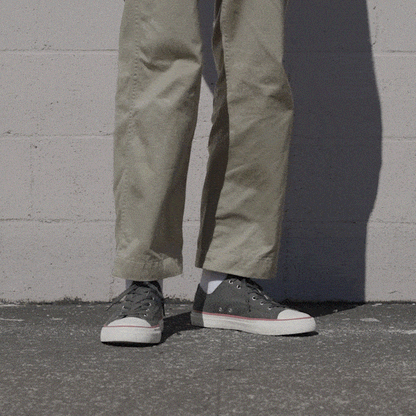 Person wearing black low-top sneakers and khaki pants shifting stance against a stone wall.