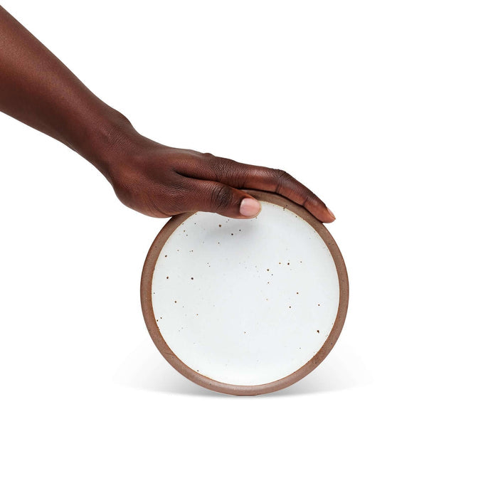A hand holding a dessert sized ceramic plate in a cool white color featuring iron speckles and an unglazed rim.