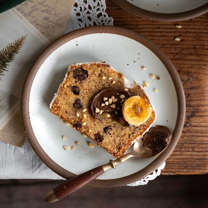 Slice of banana bread with raisins, chocolate spread, banana slice, and chopped nuts on a cool white ceramic dessert plate with a wooden-handled spoon.