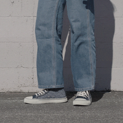 Person wearing navy low-top sneakers and blue jeans shifting stance against a stone wall.