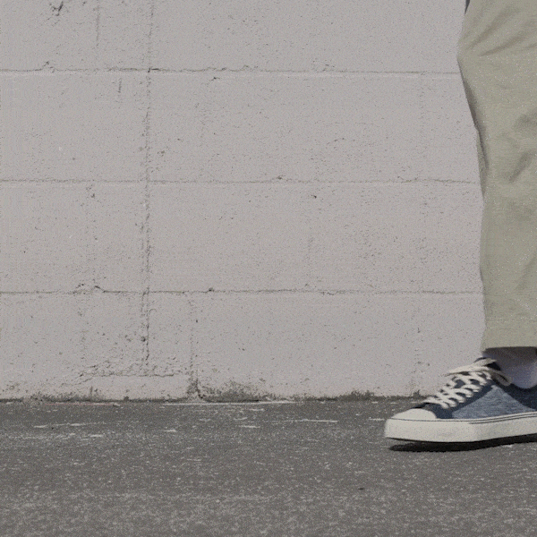 Person wearing navy low-top sneakers and khaki pants shifting stance against a stone wall.