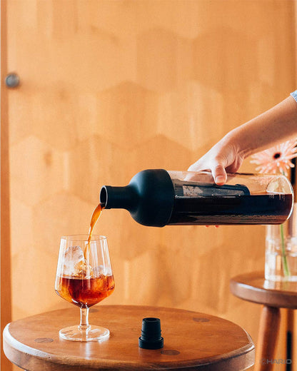 Person pouring dark liquid from a bottle into a glass on a wooden surface.
