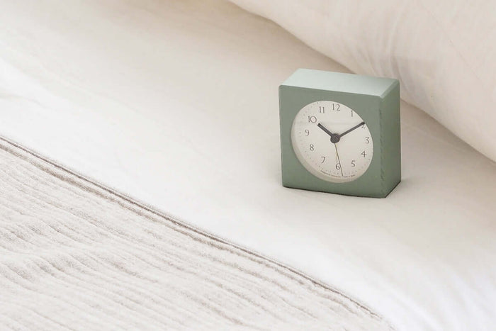 Square light green clock on a bed with white bedding