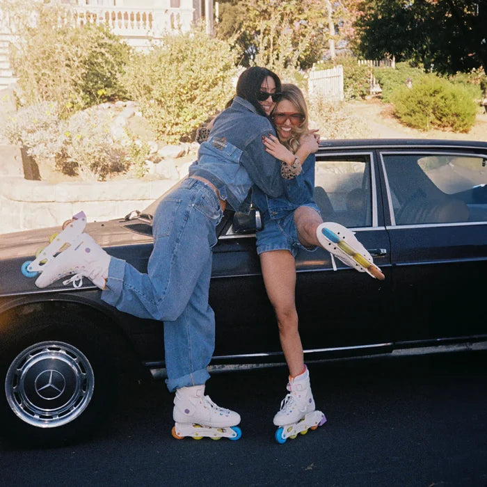 Two women on roller skates hugging in front of a black car with a Mercedes logo.
